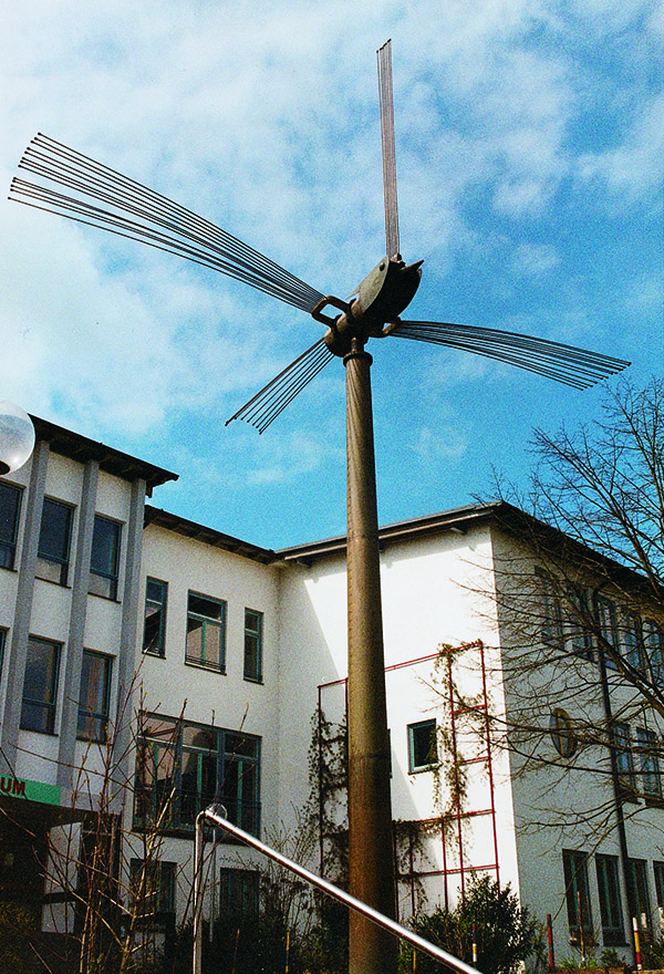 Das Foto zeigt einen Stahl-Vogel auf einer Säule. Seine Federn sind aus langen Stangen, die bei Wind klingen.