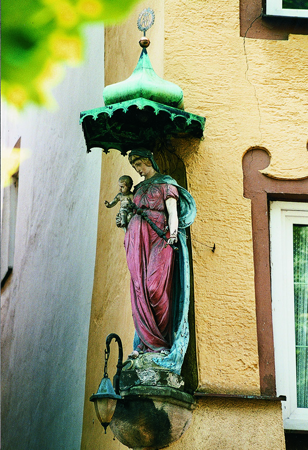 Das Foto zeigt die Statue Maria mit Kind an einer Häuserwand. Maria hat ein rotes Kleid an.