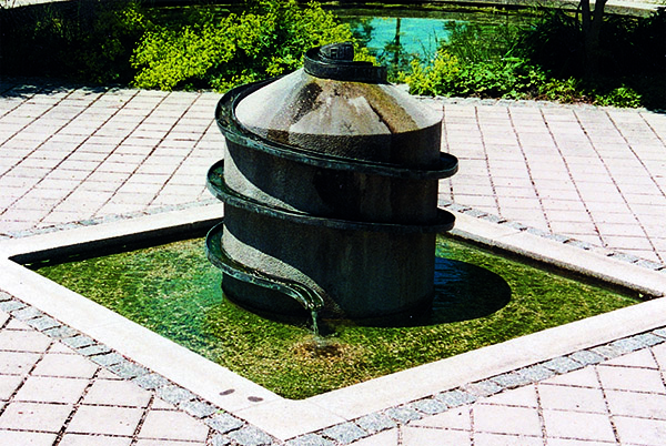 Das Foto zeigt einen Brunnen aus Metall um dessen Mitte sich eine Schlange windet. Hieran fließt das Wasser herunter.