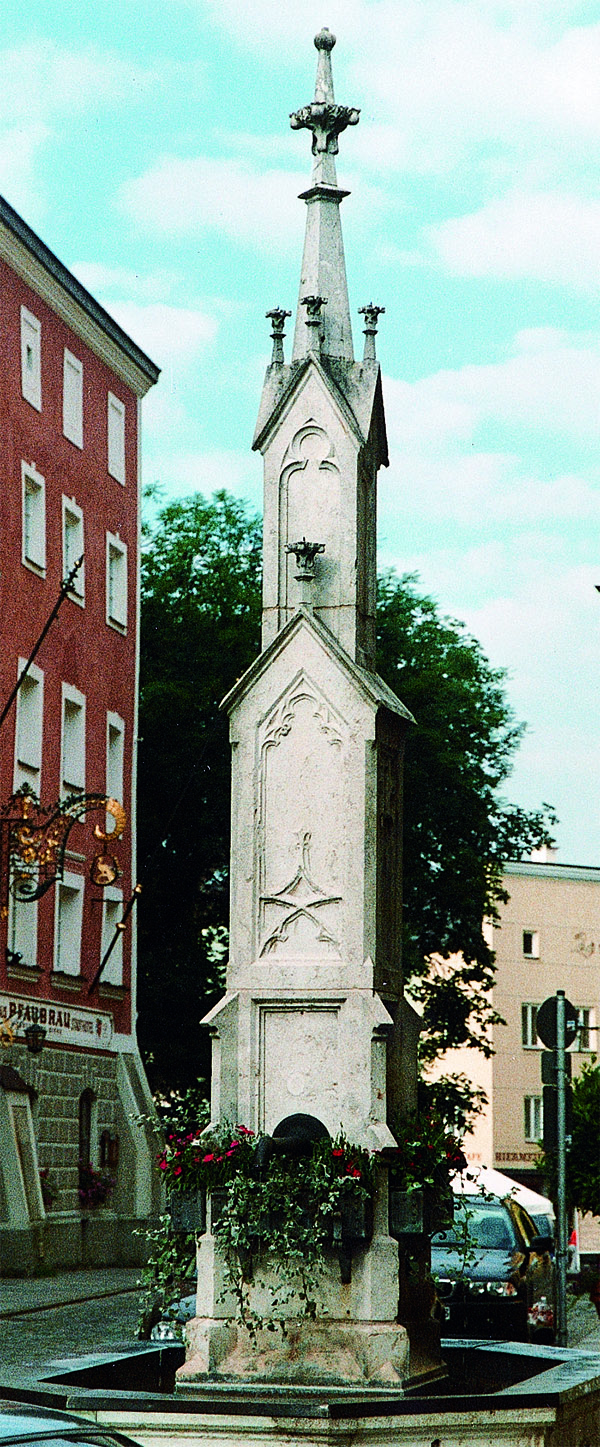 das Foto zeigt einen Steinbrunnen in der Hauptstraße. Eine große Säule ragt in der Mitte in die Höhe.