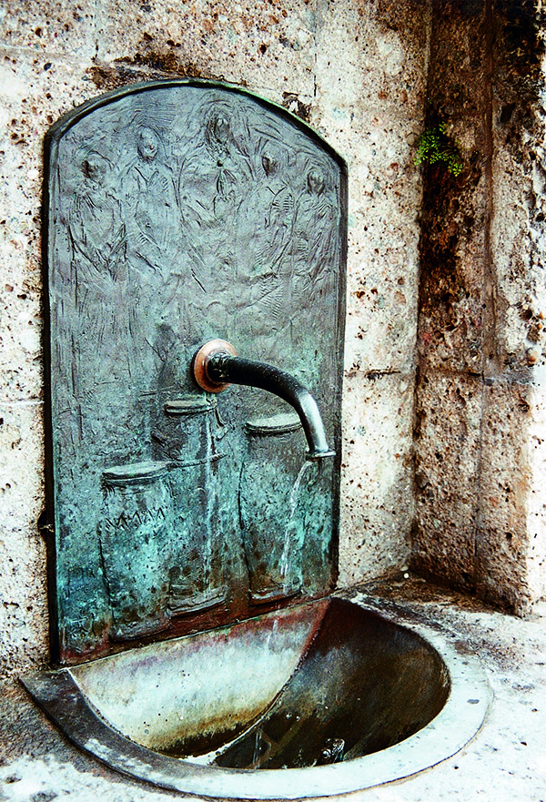 Das Foto zeigt einen Wandbrunnen mit dem Gleichnis der Hochzeit zu Kana im Hintergrund.