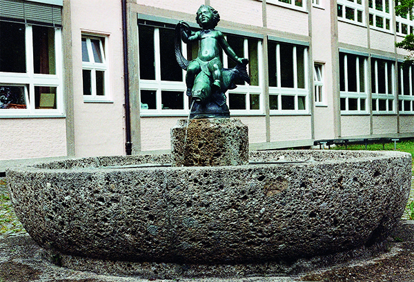 Das Foto zeigt einen Steinbrunnen, in dessen Mitte ein Junge auf einem Fisch aus Bronze, sitzt