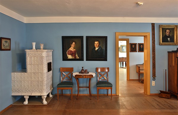 Detailansicht des Helene-Sedlmayr-Zimmers in dem unter anderem ein weisser Kachelofen, sowie zwei Bilder zu sehen sind.